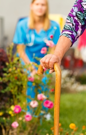In the foreground, a senior patients arm holding a cane. In the background, there is a garden and a doctor watching the senior