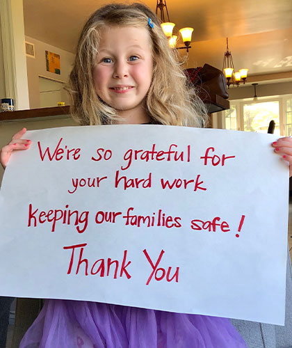 A girl in a purple dress holding a sign that reads Were so grateful for your hard work Keeping our families safe! Thank you!