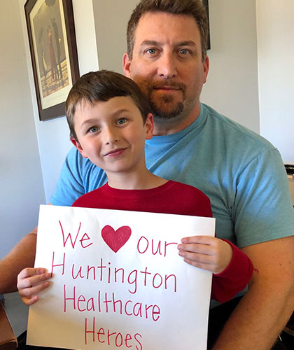 A father and son. The son holds a sign that reads We love our Huntington Healthcare heroes.