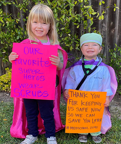 Two children waring physicain costumes holding signs. One sign reads Our favorite super heros wear scrubs. A second reads Thank you for keeping us safe now so we can save you later.