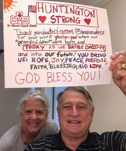 A smiling couple. A man holds a sign that reads Huntington Strong. Thank you doctors nurses, 1st responders for your work yester-year when our grandchildren were born and today as we battle COVID-19 and into our future! You bring us: hope, joy, peace, purpose, faith, blessing,and life. God Bless You!
