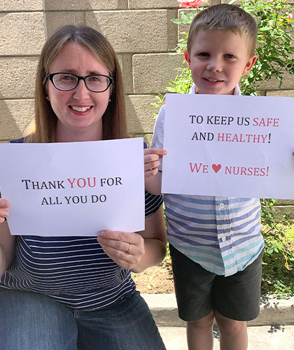 A mother and son. The mother holds a sign that reads Thank you for all you do. The son holds a sign that reads To keep us safe and healthy! We love Nurses.