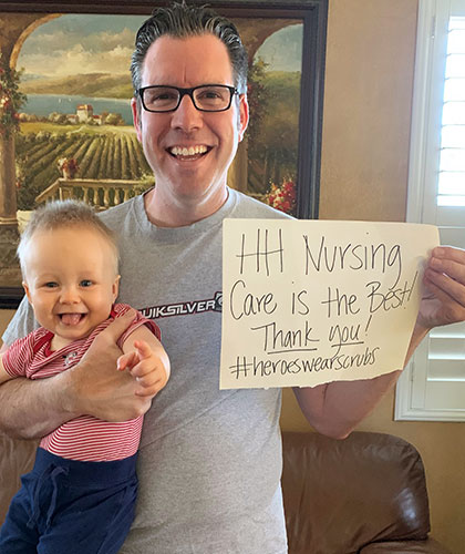 A father holding his baby son in one arm and a sign in the other that reads HH Nursing Care is the Best Thank You!