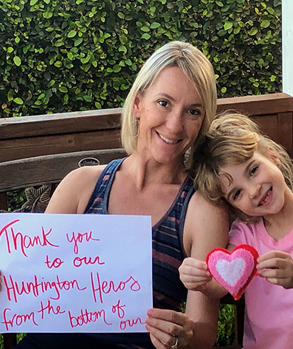 A mother and daughter. The daughter is holding a hert shaped craft. The mother is hodling a sign that reads Thank you to our Huntington hearos from the bottom of our heart.