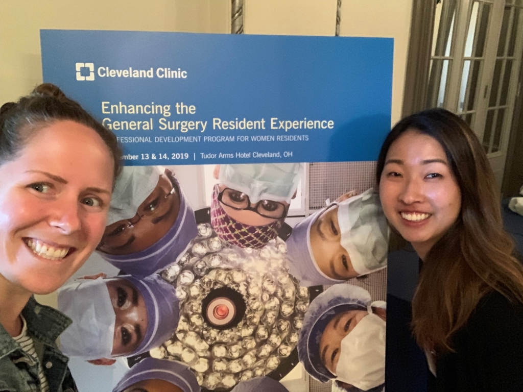 Two residents standing in front of a panel graphic that reads Enhancing the General Surgery Resident Experience