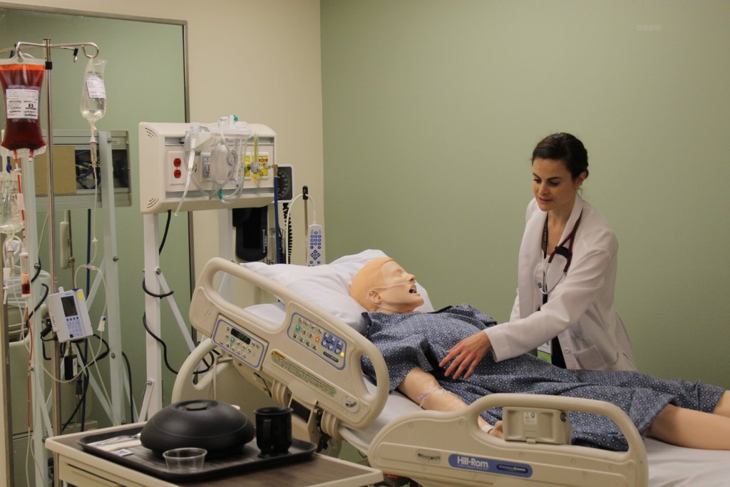 A resident checks on a simulated patient dummy that is laying on a hospital bed