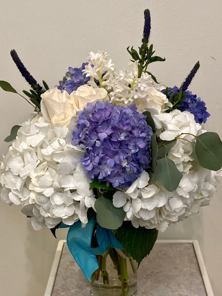 A bunch of purple and white flowers tied with. Blue ribbon in a clear vase