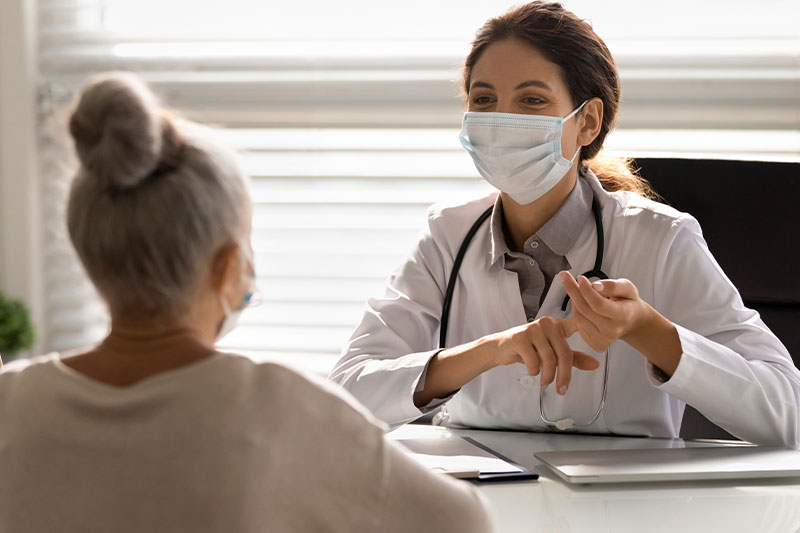 Young female doctor with mask talks to older woman patient