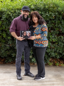Husband and wife smiling with photo of twin daughters.