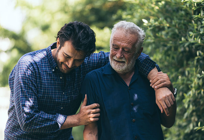 A son with his arm around his father, walking outside by trees