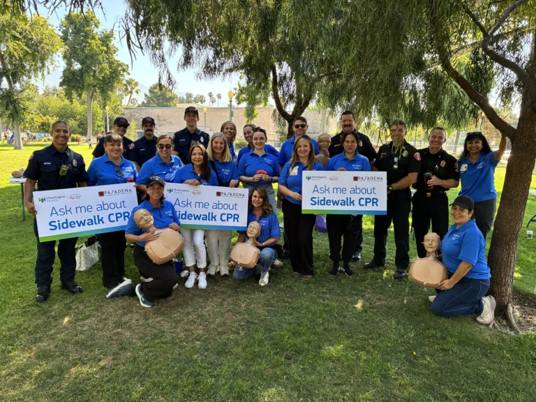 Group photo of Huntington Health team members, Pasadena Fire Department personnel and community partners gathered in a park for a Sidewalk CPR event. Several participants hold signs reading “Ask me about Sidewalk CPR,” while others hold CPR training manikins used to teach hands-only CPR. The group stands beneath large shade trees on a sunny day, highlighting a collaborative community effort to promote lifesaving CPR education.