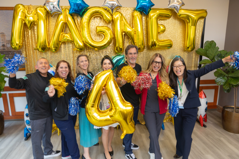 Group of eight adults standing indoors in front of a gold glitter backdrop decorated with large gold balloon letters spelling “MAGNET” and star-shaped balloons. The group holds blue, gold, and red pom-poms and a large gold balloon shaped like the number 4, celebrating Huntington’s fourth Magnet redesignation.